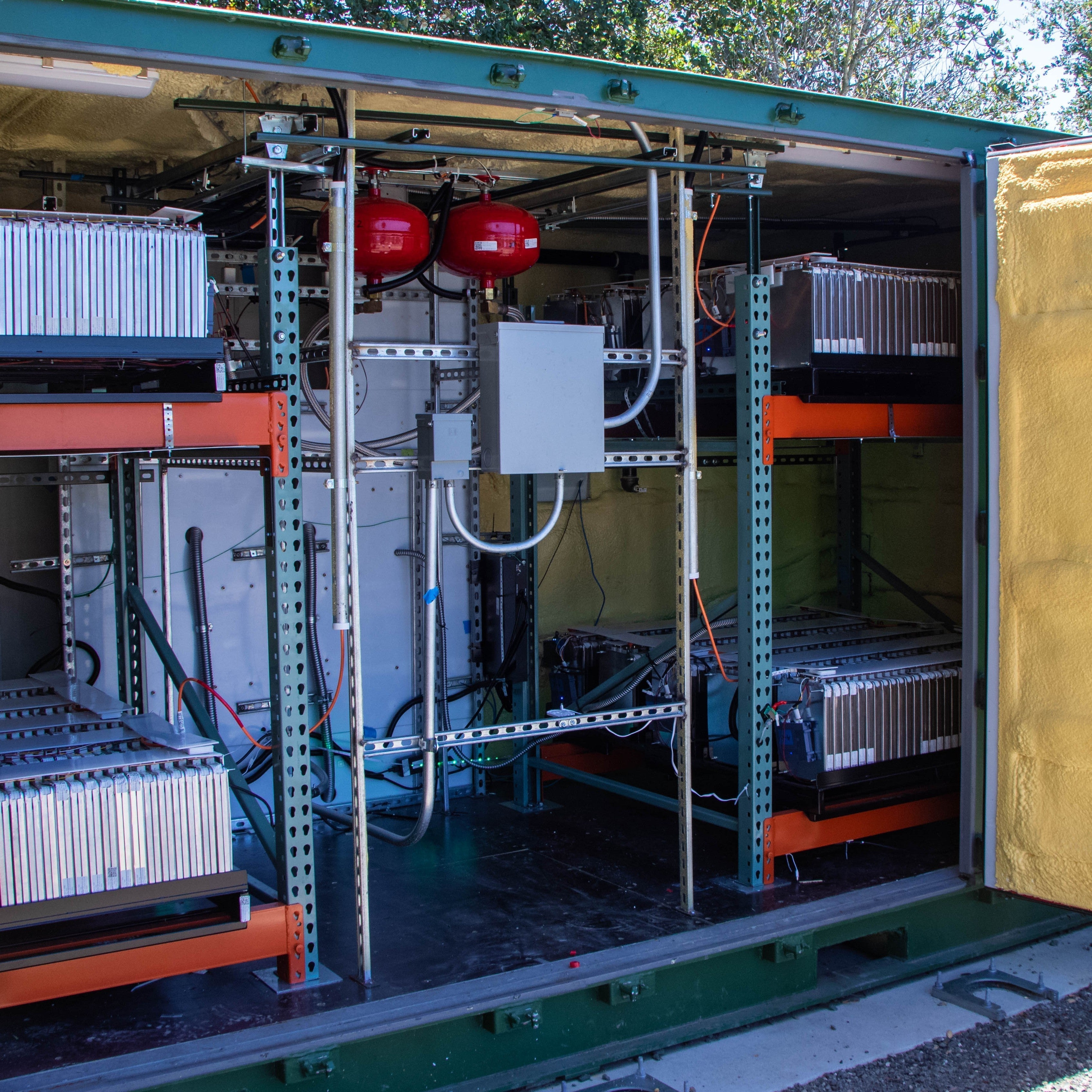Open green utility container with yellow insulation on the door, showing internal racks, cables, and equipment in an outdoor setting.